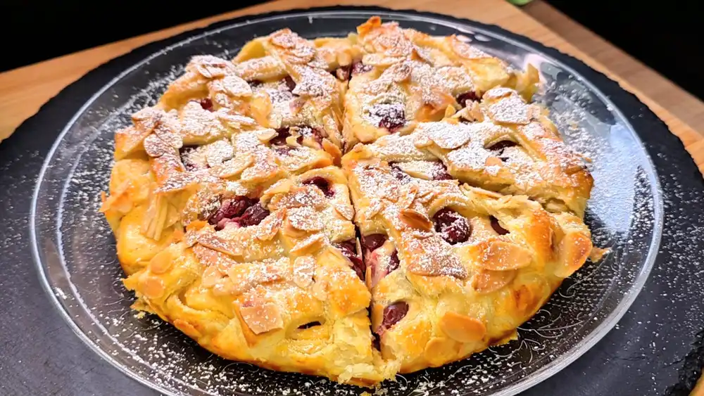 Goldbraun gebackener, runder Blätterteig-Kuchen mit einem eleganten Teiggitter und gehobelten Mandeln, gefüllt mit Quarkcreme und roten Sauerkirschen auf einer Kuchenplatte.
