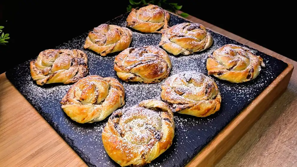 Goldbraun gebackene, gedrehte Blätterteigschnecken mit sichtbarer, leicht karamellisierter Schoko-Schaumkuss-Füllung, bestäubt mit Puderzucker.