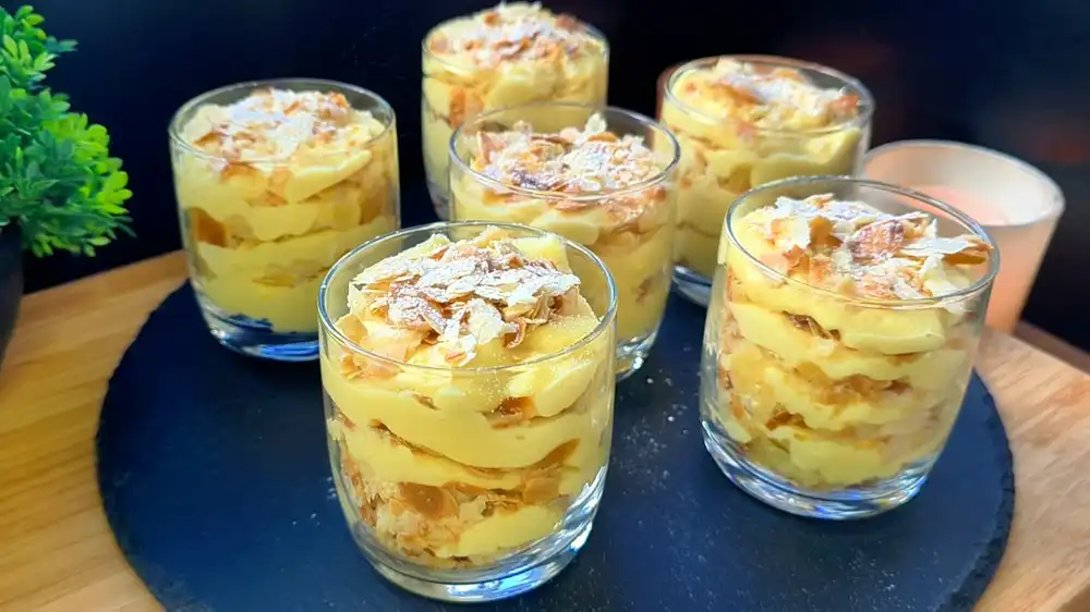 Drei elegant angerichtete Dessertgläser mit dem Vanille-Blätterteig Schichtdessert, abwechselnd gefüllt mit gelber Vanille-Mascarpone-Creme und krossen Mandel-Blätterteig-Teigkrümeln, fein mit Puderzucker bestäubt.