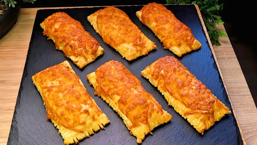 Goldbraun gebackene Blätterteig-Würstchen-Stangen mit einer krossen Kruste aus geschmolzenem Käse, angerichtet auf einer Picknickdecke.