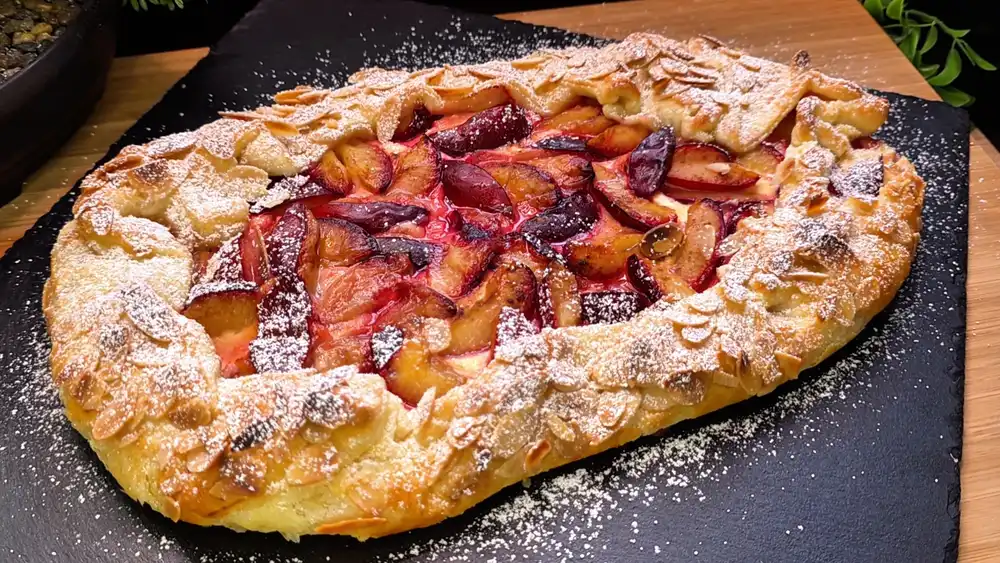 Eine goldbraun gebackene, rustikale Blätterteig-Zwetschgen-Galette mit Quarkcreme und Mandelblättchen auf Backpapier, fein mit Puderzucker bestäubt.