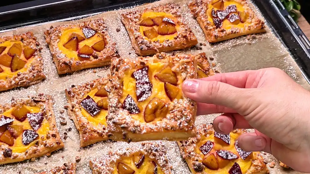 Detailansicht eines einzelnen Blätterteig-Teilchens mit Fokus auf die glänzende Puddingfüllung, die weich gebackene Zwetschge und den krossen Haselnussrand mit Puderzucker.