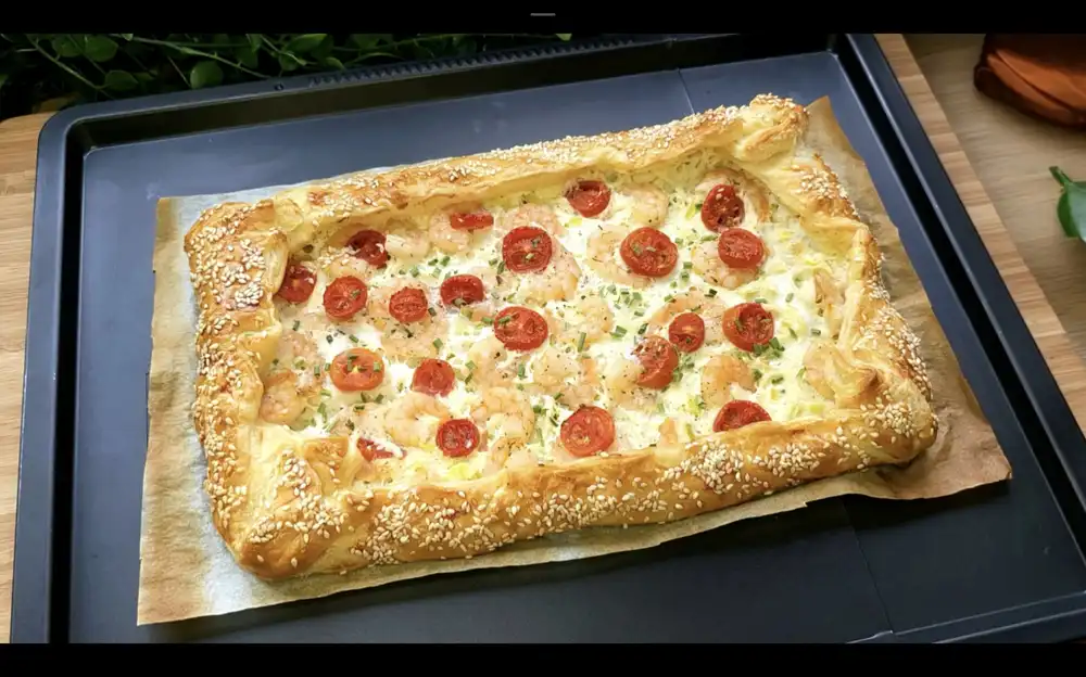 Eine goldbraun gebackene, rustikale Blätterteig-Galette gefüllt mit cremiger Frischkäse-Masse, rosa Garnelen, roten Tomatenscheiben und Sesam auf einem rustikalen Holzbrett.