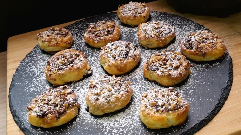 Mehrere goldbraun gebackene Blätterteig-Schnecken mit Schokoladenfüllung, Preiselbeeren und gehackten Haselnüssen, fein mit Puderzucker bestreut auf einer Etagere.