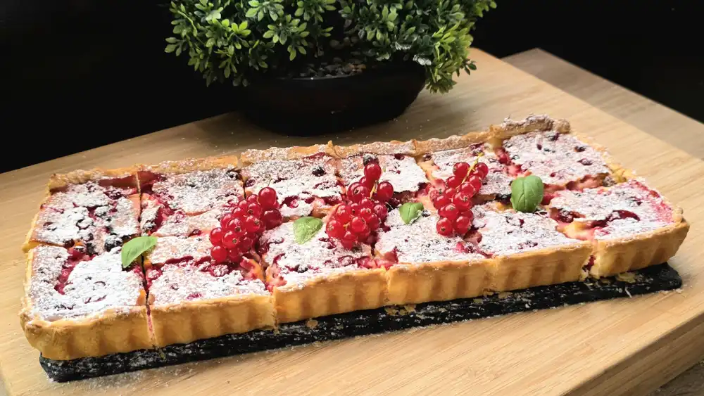 Ein goldbraun gebackener Käsekuchen aus Blätterteig in einer Backform, üppig gefüllt mit gelber Vanille-Quark-Masse und roten Johannisbeeren, fein mit Puderzucker bestäubt.