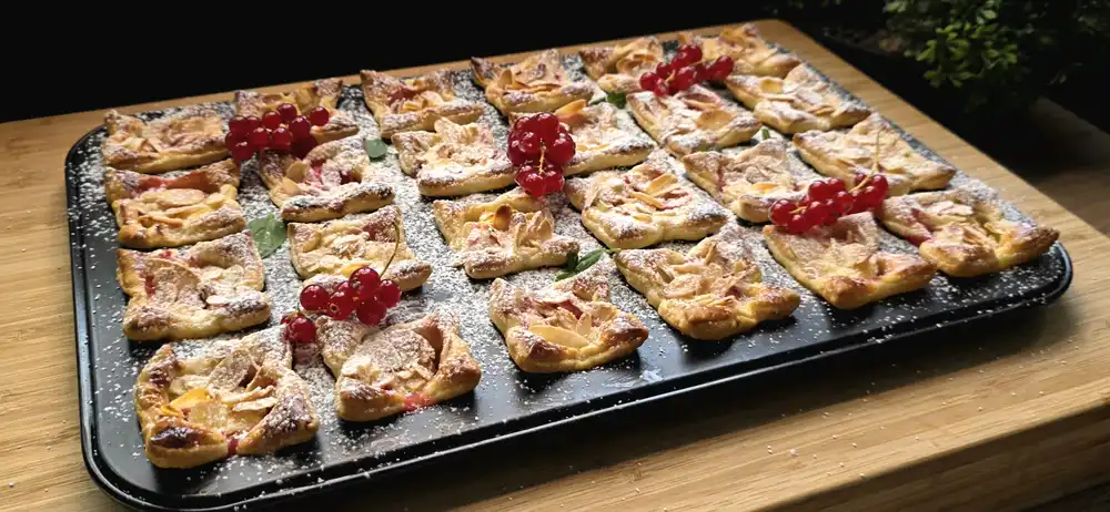 Eine Muffin-Form voller goldbraun gebackener Blätterteig-Mini-Muffins mit gelber Quarkfüllung, roten Johannisbeeren und gehobelten Mandeln, fein mit Puderzucker bestäubt.