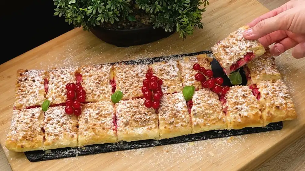 Eine große, goldbraun gebackene Blätterteig-Quarktasche mit roten Johannisbeeren und gehackten Mandeln, fertig in Stücke geschnitten auf einer Servierplatte.