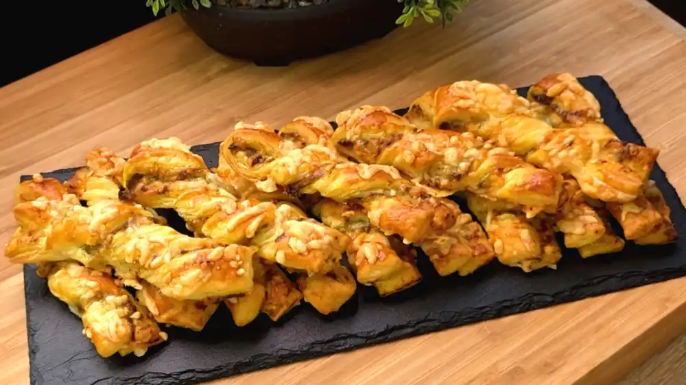Mehrere knusprig gedrehte Blätterteigstangen mit grünem Pesto und einer goldbraun überbackenen Käseschicht auf einem Kuchengitter.