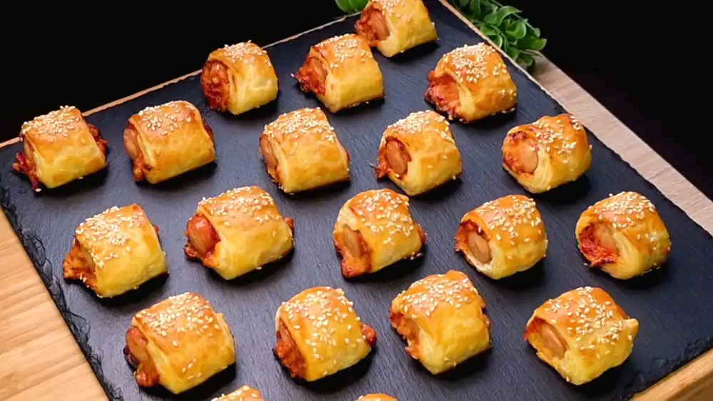 Viele goldbraun gebackene, mundgerechte Blätterteig-Schnecken mit Sesam-Topping, gefüllt mit Pizzasoße, geschmolzenem Käse und einem Würstchen in der Mitte.