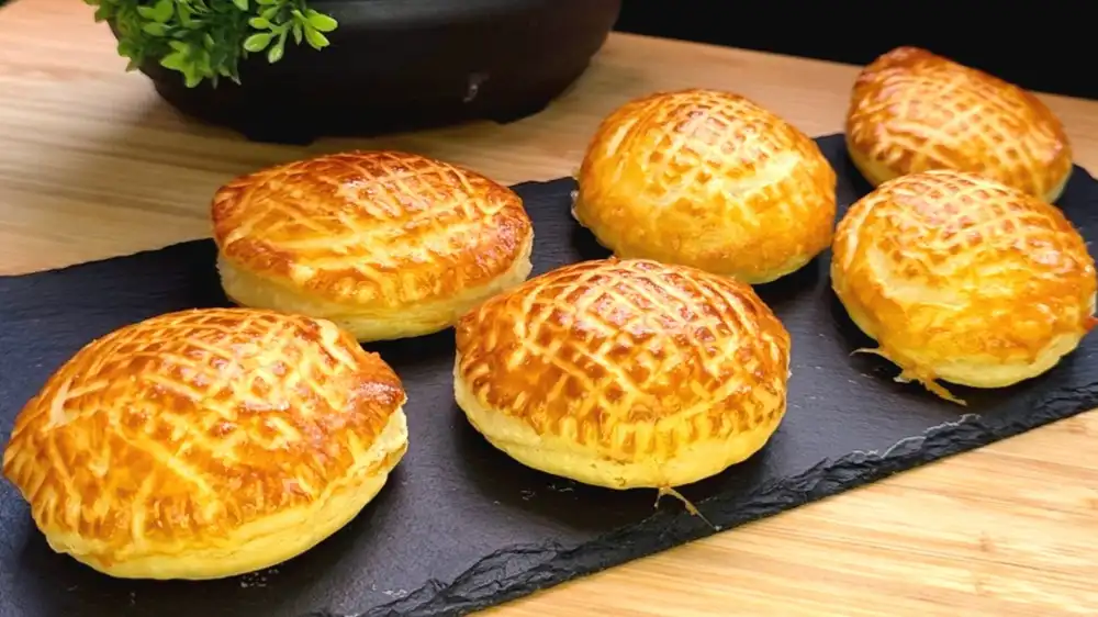 Vier goldbraun gebackene, runde Käsetaschen aus Blätterteig mit einem feinen Gabelmuster auf der Oberfläche, angerichtet auf einem rustikalen Holzbrett.