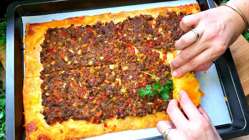 Ein großes, goldbraun gebackenes Lahmacun aus Blätterteig, reichlich belegt mit würzigem Hackfleisch, Paprika und frischer Petersilie auf einem Holzbrett.