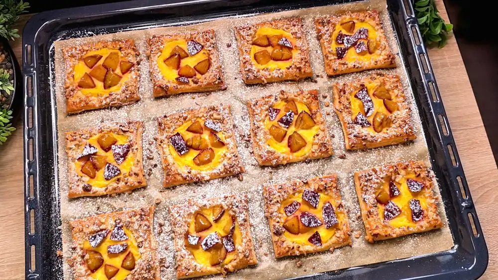 Zwölf goldbraun gebackene, eckige Blätterteig-Teilchen, gefüllt mit gelbem Vanillepudding, belegt mit violetten Zwetschgenspalten und mit gehackten Haselnüssen bestreut.