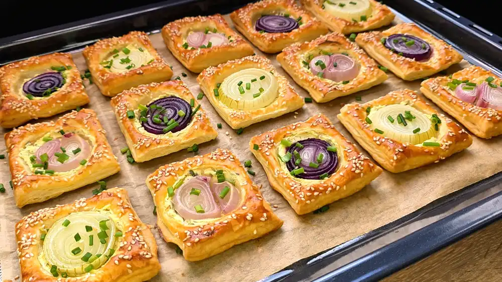 Eine Platte mit 15 goldbraun gebackenen, eckigen Blätterteig-Zwiebelkuchen, bestrichen mit Kräuterfrischkäse, belegt mit roten Zwiebelringen und mit Sesam bestreut.