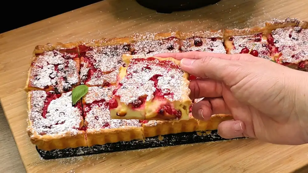 Detailansicht eines angeschnittenen Stücks Käsekuchen mit Fokus auf den knusprigen Blätterteigrand, die cremige Quarkfüllung und die saftig aufgesprungenen roten Johannisbeeren.