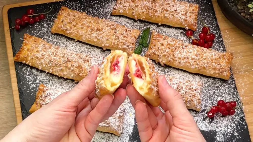 Detailansicht einer angeschnittenen Blätterteigstange mit Fokus auf die samtige Vanille-Quarkfüllung, die aufgeplatzten roten Johannisbeeren und den knusprigen Mandel-Rand mit Puderzucker.