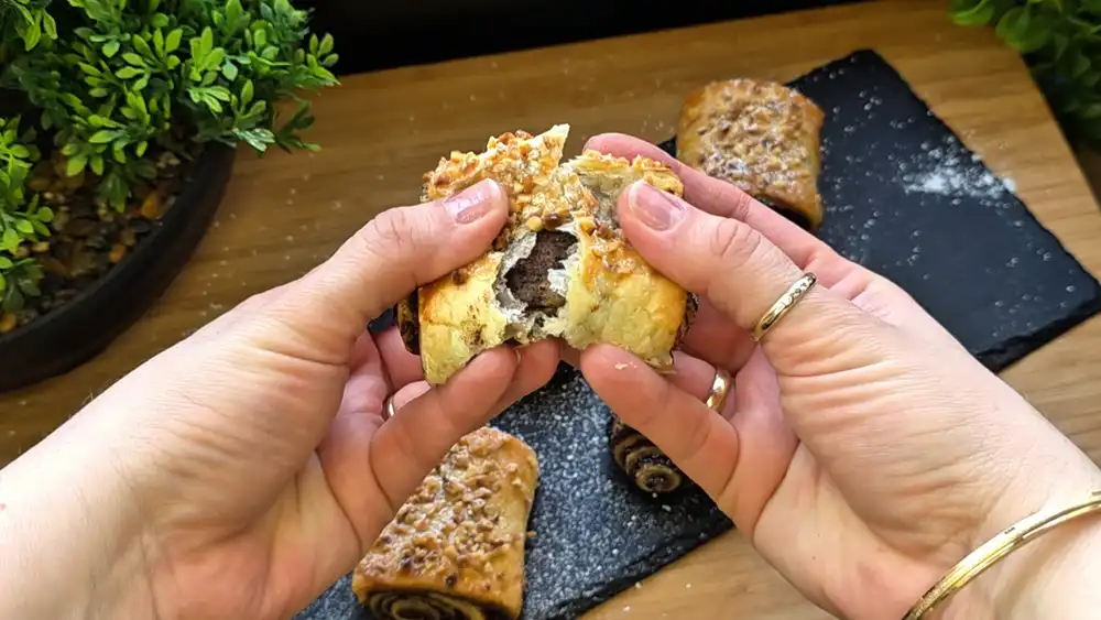Detailansicht einer frisch gebackenen Kakao-Nuss-Rolle aus Blätterteig mit Fokus auf die krossen Teigschichten und die heraustretende Kakaofüllung mit gerösteten Nüssen.