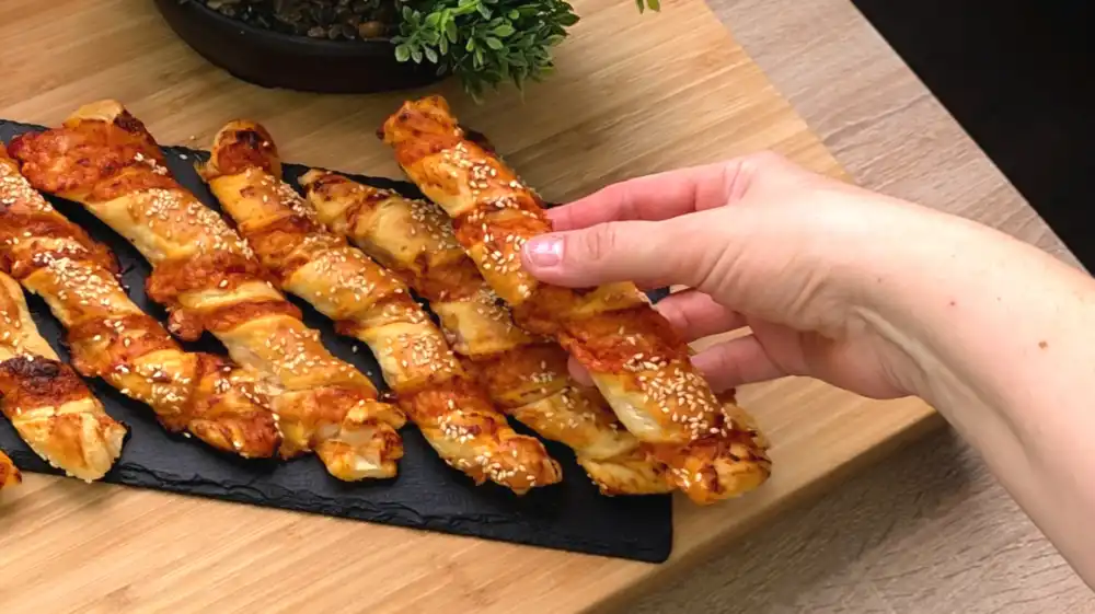 Detailansicht einer knusprig gedrehten Blätterteigstange mit Fokus auf die sichtbare rote Tomatensauce, den krossen Käse und die gerösteten Sesamsamen.