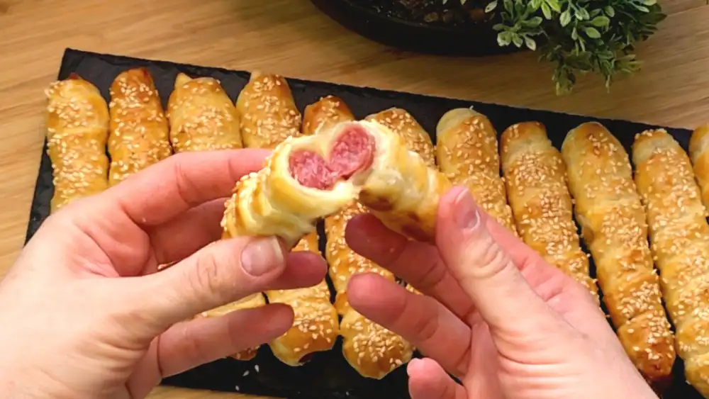 Detailansicht eines frisch gebackenen Salami-Blätterteig-Stäbchens mit Fokus auf die knusprigen Teigschichten, den goldgelben Glanz und die gerösteten Sesamsamen.