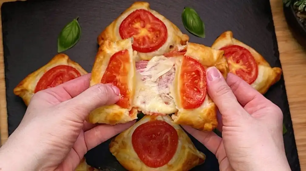 Detailansicht einer frisch gebackenen Blätterteigtasche mit Fokus auf die saftige Tomate obenauf, den austretenden geschmolzenen Mozzarella und die knusprigen Teigschichten.