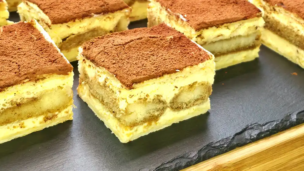 Detailansicht einer angeschnittenen Tiramisu-Schnitte mit Fokus auf die knusprigen Blätterteigplatten, die Vanille-Mascarpone-Creme und die kaffeegetränkten Löffelbiskuits.