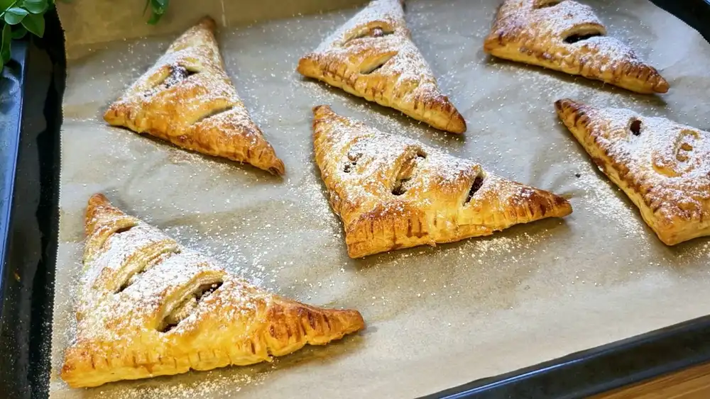 Sechs goldbraun gebackene Blätterteig-Dreiecke mit Marzipan und Zwetschgen, fein mit Puderzucker bestäubt auf einem Kuchengitter.