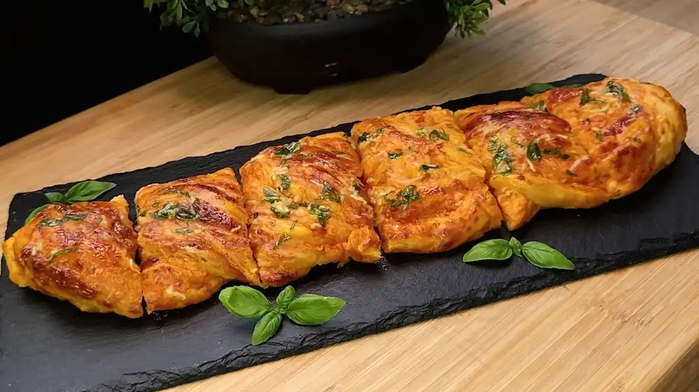 Eine rustikale Servierplatte mit einer goldbraun gebackenen Blätterteig-Pizza-Babka, die mit glänzendem Knoblauch-Basilikum-Öl bestrichen ist.