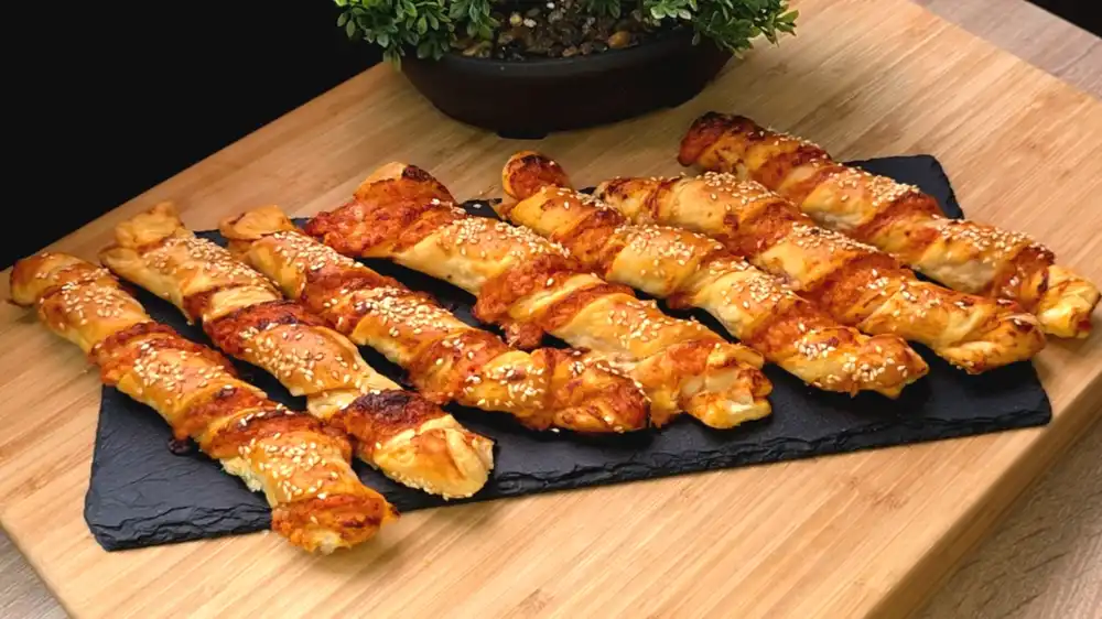 Mehrere goldbraun gebackene, gedrehte Blätterteigstangen mit Sesam-Topping, aus denen geschmolzener Käse und rote Pizzasauce hervorlugen.