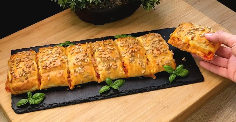 Ein goldbraun gebackenes, großes Pizza-Kissen aus Blätterteig, bestreut mit knusprigen Sonnenblumenkernen, aufgeschnitten auf einem rustikalen Holzbrett.