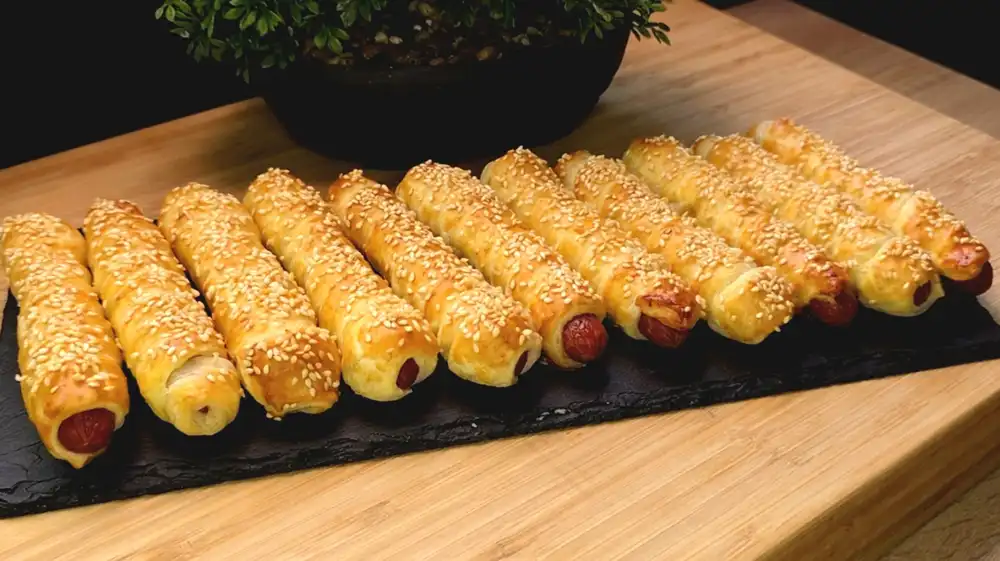 Zwölf goldbraun gebackene, mit Sesam bestreute Blätterteig-Stangen, aus denen an den Enden krosse Mini-Salamis herausschauen, serviert in einem Glas auf einem Sommertisch.