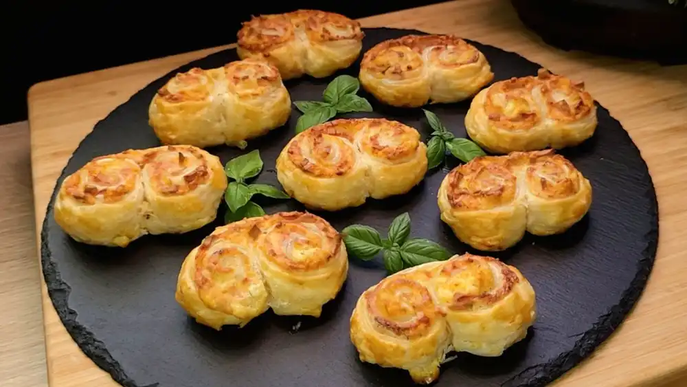 Eine weiße Servierplatte voller goldbraun gebackener Blätterteig-Schweineohren mit geschmolzenem Käse und Schinkenstückchen.