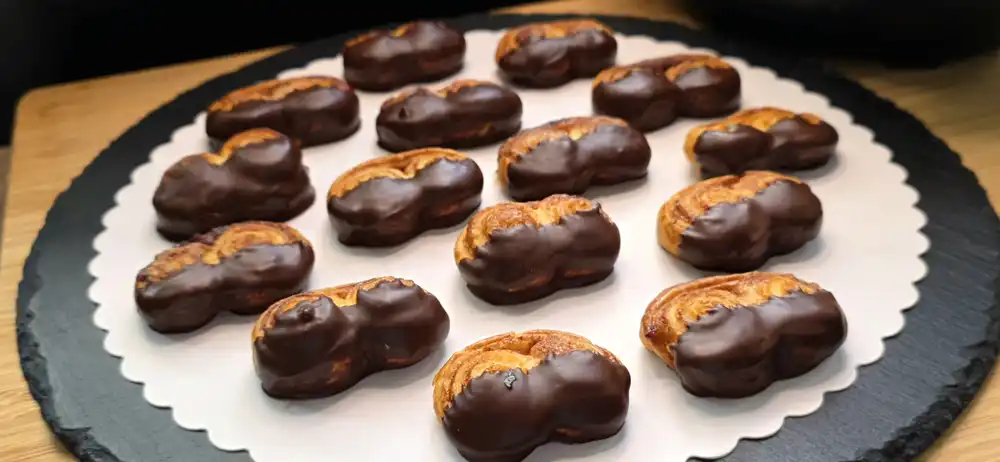Viele frisch gebackene, goldbraune Schweineöhrchen aus Blätterteig mit einer dunklen Schokoladenseite, angerichtet auf einer Etagere.