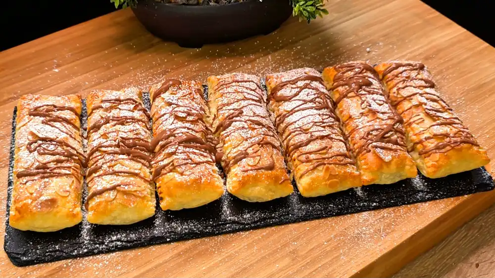 Eine stilvolle Servierplatte mit goldbraun gebackenen Blätterteig-Schoko-Quark-Riegeln, dekoriert mit gehobelten Mandeln, Puderzucker und feinen Schokostreifen.