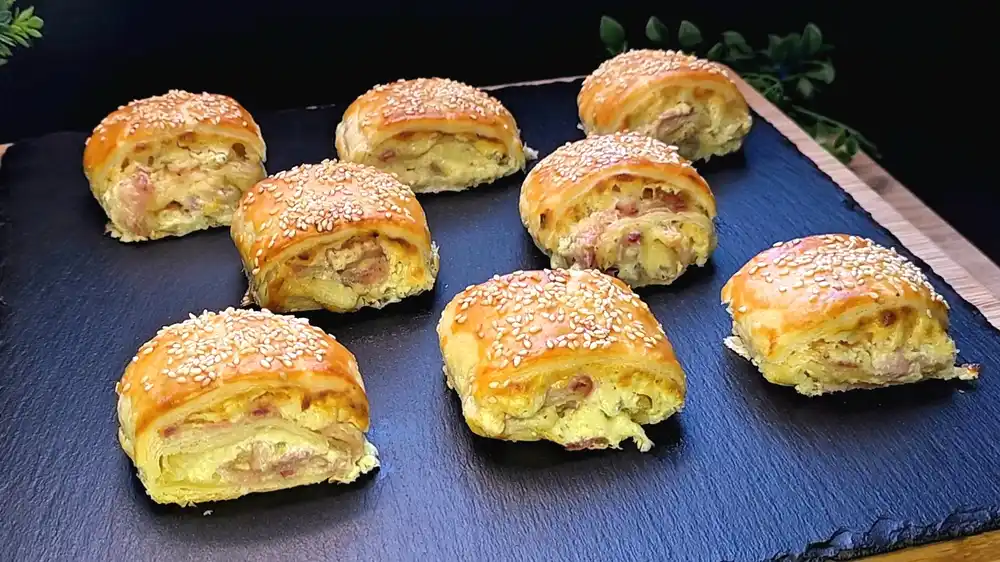 Acht frisch gebackene, eckig-runde Blätterteig-Schnecken mit goldbrauner Kruste, gefüllt mit cremigem Kräuterfrischkäse, geschmolzenem gelbem Käse, krossen Speckwürfeln und mit Sesam bestreut.