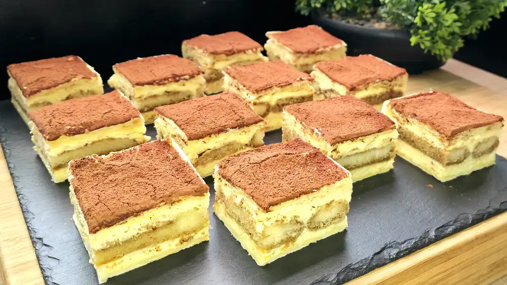 Mehrere elegant aufgeschichtete Tiramisu-Schnitten aus Blätterteig auf einer Servierplatte, üppig mit ungesüßtem Kakaopulver bestäubt.