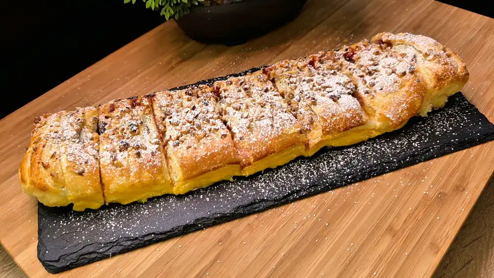 Ein goldbraun gebackener, krosser Blätterteig-Zimt-Strudel mit sichtbaren Einschnitten, gehackten Haselnüssen und Puderzucker auf einer Kaffeetafel.
