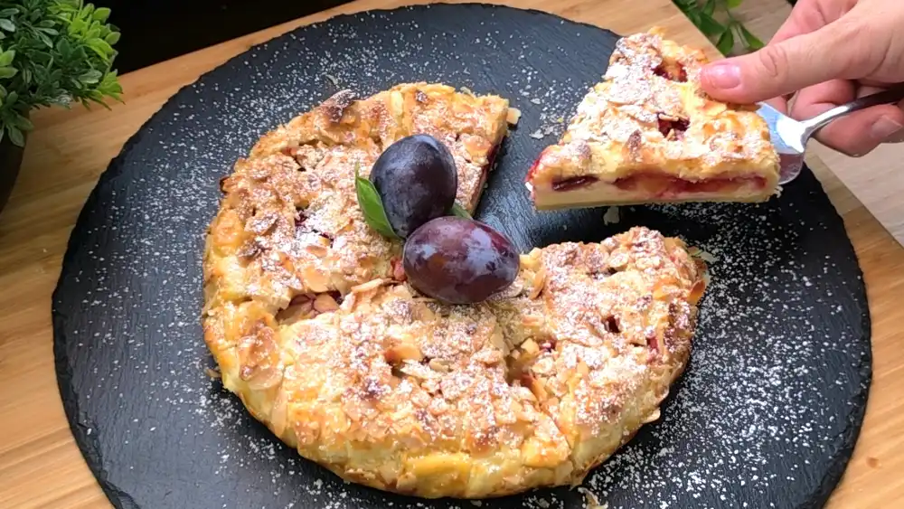 Ein runder, goldbraun gebackener Blätterteig-Kuchen mit einem dekorativen Gittermuster, gefüllt mit weißer Quarkcreme, lila Zwetschgen und gehobelten Mandeln auf einer Tortenplatte.