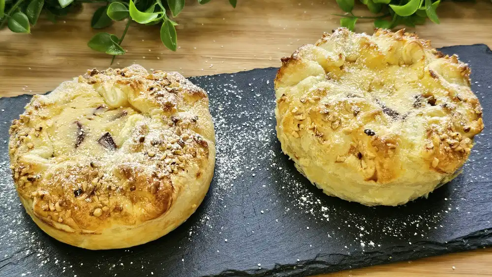Zwei goldbraun gebackene Mini-Zwetschgen-Käsekuchen aus Blätterteig mit gehackten Haselnüssen und Puderzucker auf einer eleganten Etagere.