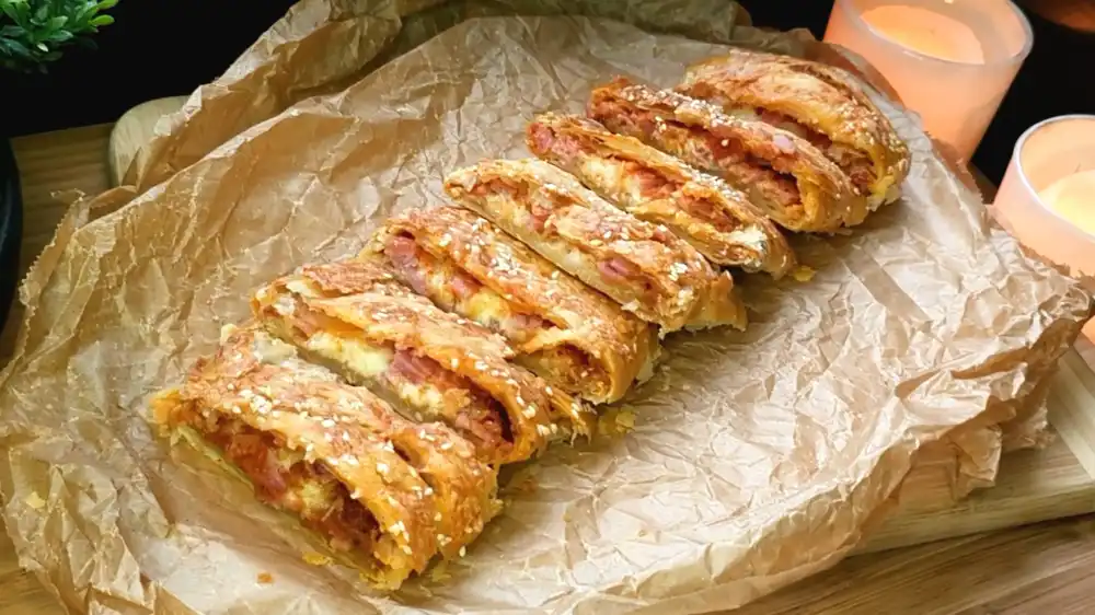 Ein goldbraun gebackener Blätterteig-Pizza-Strudel mit geschmolzenem Käse, Sesam-Bestreuung und feinen Schinkenwürfeln auf einem Holzbrett.