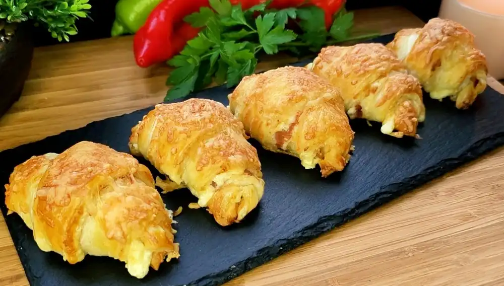 Goldbraun gebackene Blätterteig-Schinken-Käse-Croissants mit überbackener Käsekruste auf einer rustikalen Holzplatte.