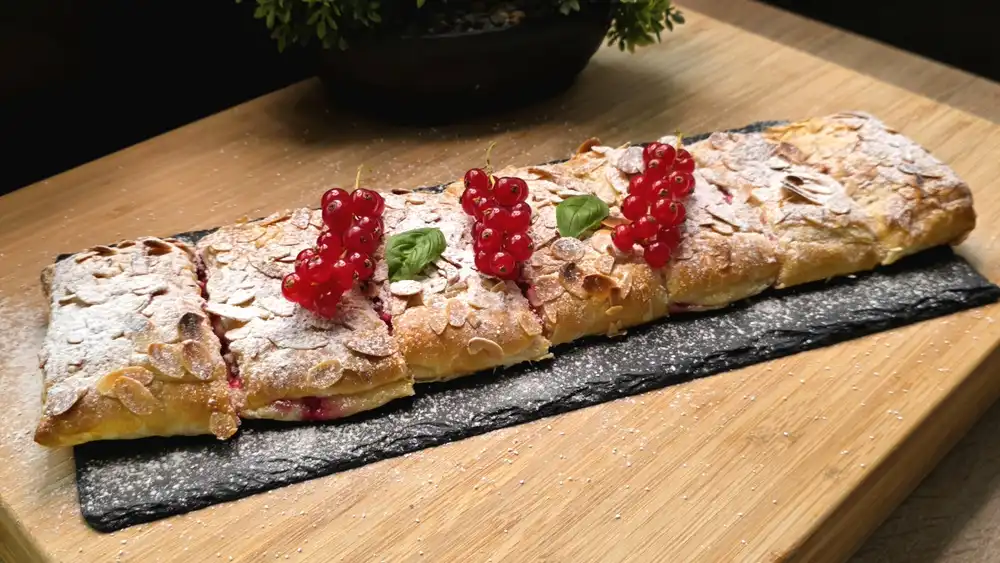 Eine goldbraun gebackene Blätterteig-Quarkrolle mit gehobelten Mandeln und Puderzucker auf einem Holzbrett, aufgeschnitten mit Blick auf die rote Beeren-Quark-Füllung.
