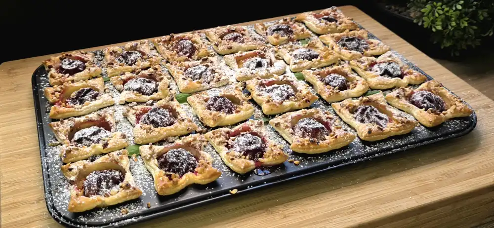 24 goldbraun gebackene Blätterteig-Mini-Muffins mit Quark, halbierten Pflaumen, gehackten Haselnüssen und Puderzucker in einem Muffinblech.