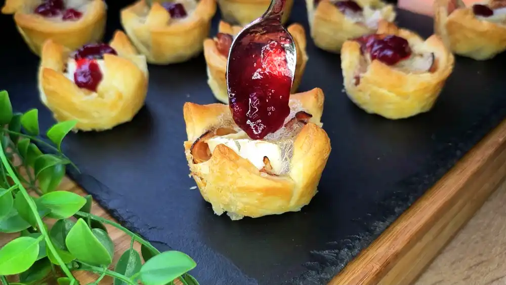 Detailansicht eines Blätterteig-Muffins mit Fokus auf den geschmolzenen Camembert, Schwarzwälder Schinken, Rosmarin und Preiselbeerklecks.