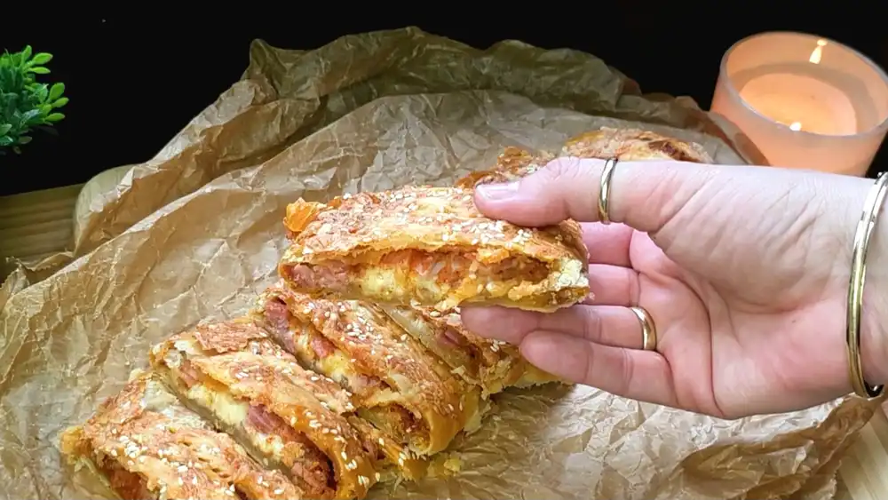 Detailansicht eines angeschnittenen Blätterteig-Pizza-Pakets mit Fokus auf die saftige Tomaten-Schinken-Käse-Füllung und die krosse Sesam-Käse-Kruste.