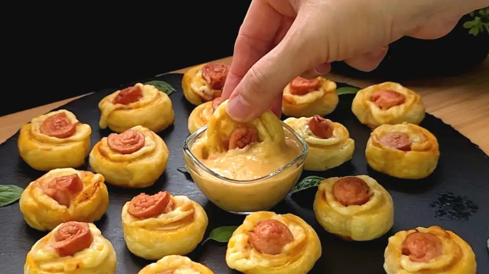 Detailansicht einzelner Mini-Würstchen-Häppchen mit Fokus auf geschmolzenen Käse, rotes Pesto und knusprigen Blätterteigmantel.