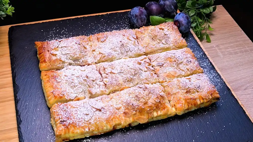 Neun goldbraun gebackene Blätterteig-Quarktaschen mit gehobelten Mandeln und Puderzucker auf einem Kuchengitter, gefüllt mit saftigen Zwetschgen.