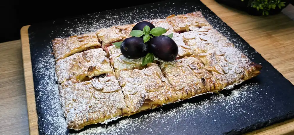 Goldbraun gebackener gedeckter Zwetschgen-Quark-Kuchen aus Blätterteig mit Mandeln und Puderzucker.